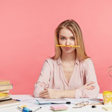 Horizontal portrait of funny female student keeps pencil on mouth, foolishes after long prepareation to pass course paper, surrounded with many books, isolated over pink background. Studying concept