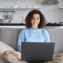 Happy hispanic teen girl using laptop computer sitting on sofa at home.